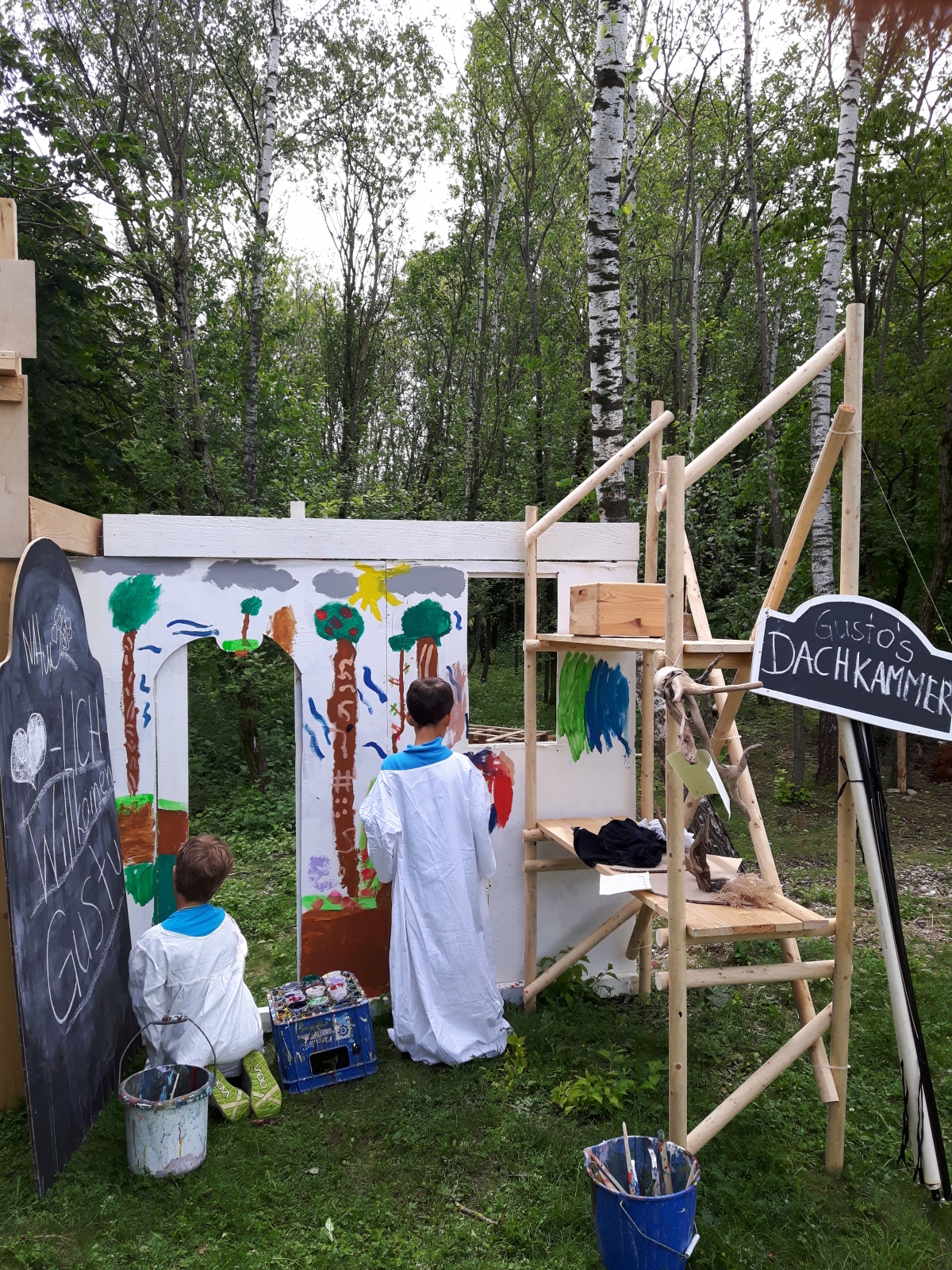 Kinder bemalen eine selbstgebaute Spiel-/Bühnenkonstruktion im Freien; rechts ein Schild „… Dachkammer“.