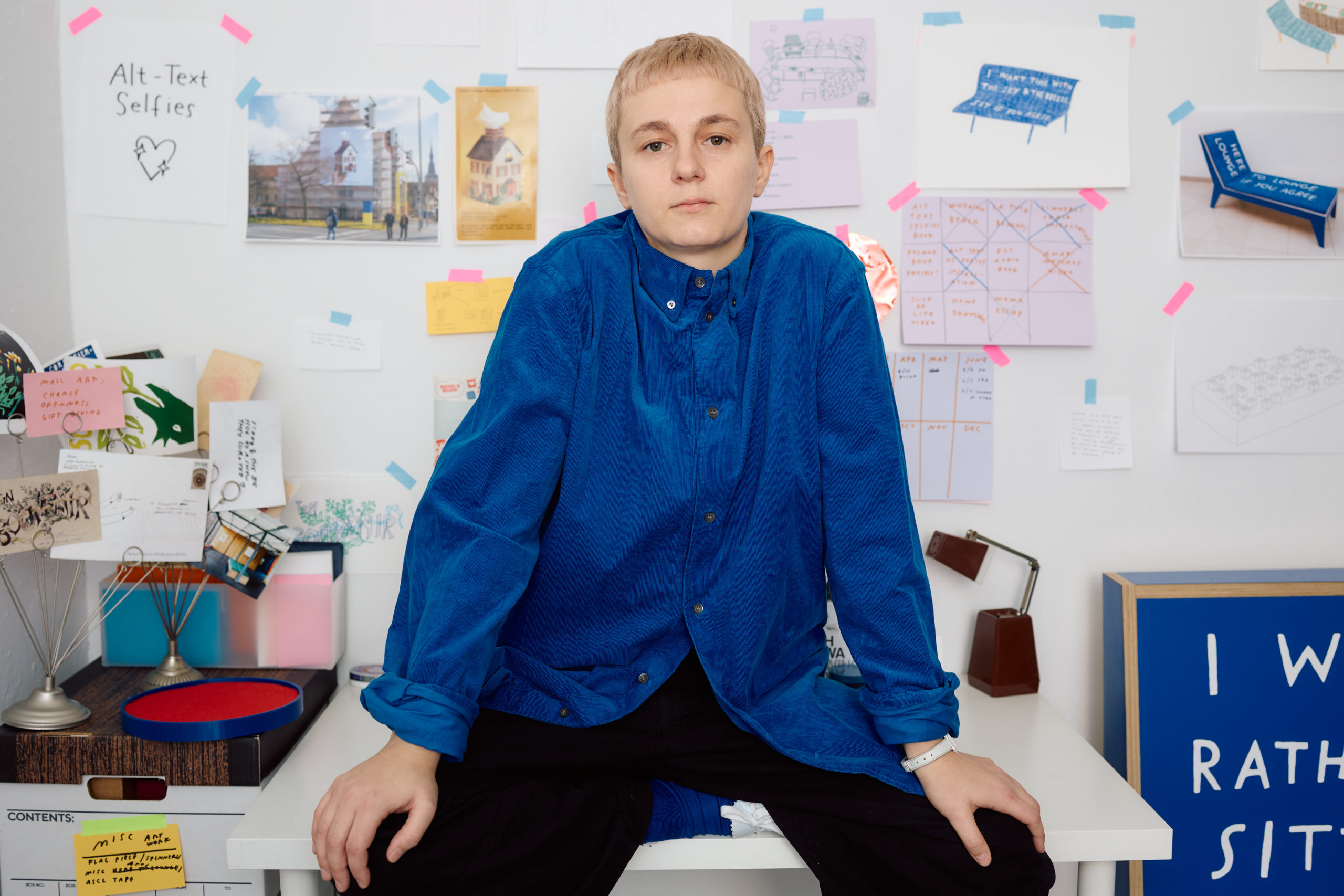 Me, a white person in a bright blue corduroy shirt and bleached blonde hair. Looking directly at the camera, relaxed but not smiling. Taped to the wall behind me are various notes about projects, sketches, and things I like. Marissa captured me looking hot and cool and boyish — what a dream!