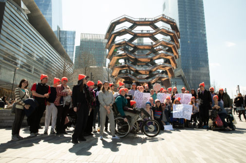 About 40 people posed in front of Thomas Heatherwick's monumental sculpture the Vessel. They are sporting neon orange, Anti-Stairs Club Lounge beanies and holding Anti-Stairs Club Lounge Signs.
